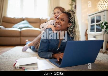 Tochter, die auf einer glücklichen Mutter lag, die zu Hause am Laptop auf dem Boden arbeitete Stockfoto