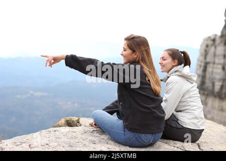 Zwei glückliche Wanderer, die auf einer Klippe sitzen und die Aussicht auf den Berg im Freien genießen und wegzeigen Stockfoto