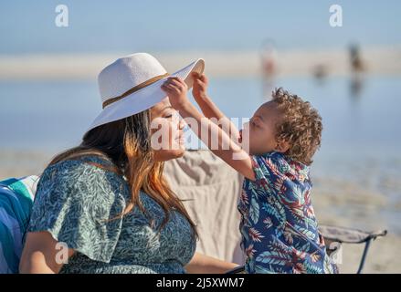 Neugieriger Sohn mit Down-Syndrom, der den Hut der Mutter am Strand hebt Stockfoto