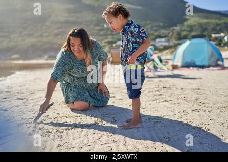 Mutter und Sohn mit Down-Syndrom zeichnen mit Stock im Sand am Strand Stockfoto