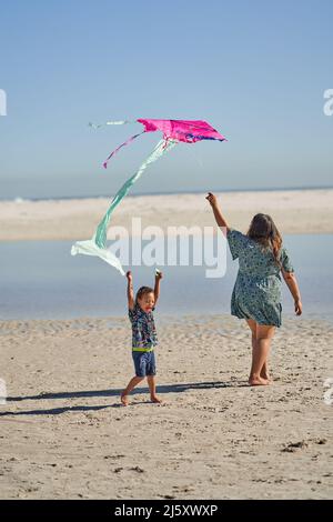 Mutter und Sohn spielen am sonnigen Strand mit Drachen Stockfoto
