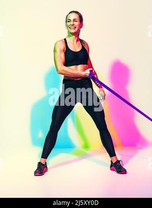 Starke Frau, die mit Widerstandsband arbeitet. Foto von Frau in schwarzer Sportkleidung auf weißem Hintergrund mit Effekt von rgb-Farben Schatten. Sport und Gesundheit Stockfoto