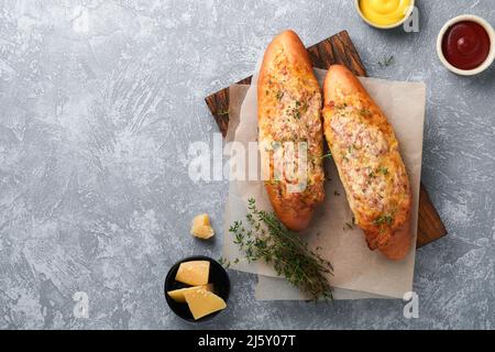 Baguette-Boote. Heiß gebackenes Sandwich auf Baguette-Brot mit Schinken, Speck, Gemüse und Käse auf Pergament und Holzständer auf grauem Beton-Hintergrund. Stockfoto