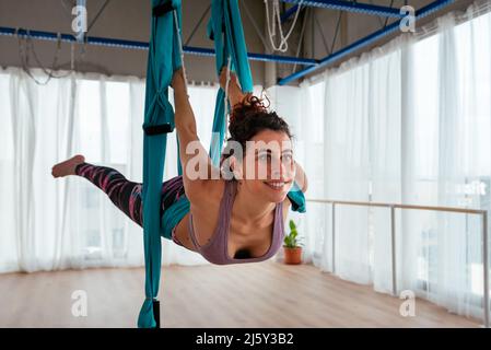 Positive aktive Frau in aktivem Ohr üben Schwan Pose auf Hängematte während Luft Yoga-Training in hellen geräumigen Studio mit Fenstern Stockfoto