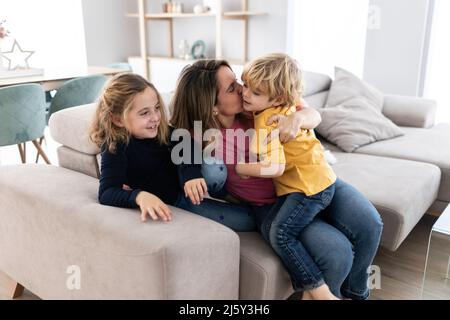 Zarte Mutter küsst niedlichen Sohn, während sie auf einer bequemen Couch mit positiver Tochter sitzt, während sie Zeit im hellen Wohnzimmer verbringt Stockfoto
