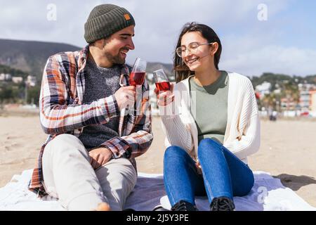 Ein positives Paar in zwangloser Atmosphäre trägt klirrende Gläser mit Rosensekt, während es an sonnigen Tagen am Sandstrand im Küstengebiet sitzt Stockfoto