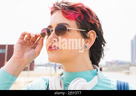 Dame mit kurz gefärbtem Haar in Sonnenbrille und Kopfhörer am Hals schaut weg, während sie die Stirn in der Nähe von Holzgeländer berührt Stockfoto