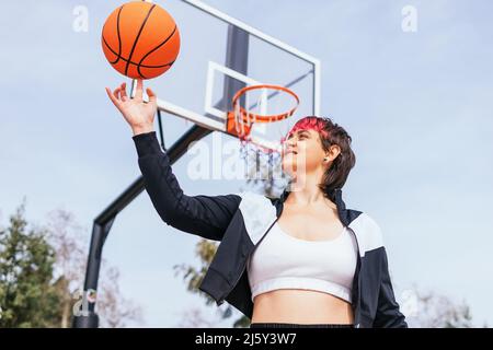 Flache Seitenansicht einer Spielerin, die den Ball in den Reifen wirft, während sie Basketball gegen den bewölkten Himmel und die grünen Bäume spielt Stockfoto