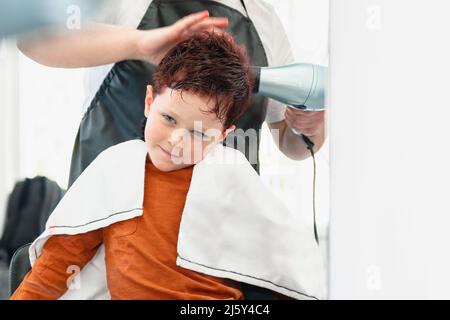 Crop anonymen Friseur in Schürze trocknen rot gefärbte Haare von niedlichen Jungen sitzen gegen den Spiegel in hellen modernen Friseursalon Stockfoto