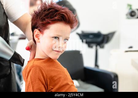 Crop anonymen Friseur in Schürze trocknen rot gefärbte Haare von niedlichen Jungen im hellen modernen Friseursalon sitzen Stockfoto