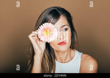 Friedliches weibliches Modell mit roten Lippen, die das Auge mit einer sanften rosa Gerbera-Blüte im Studio vor beigem Hintergrund bedecken Stockfoto