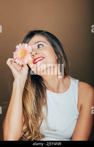 Friedliches weibliches Modell mit roten Lippen, die das Auge mit einer sanften rosa Gerbera-Blüte im Studio vor beigem Hintergrund bedecken Stockfoto