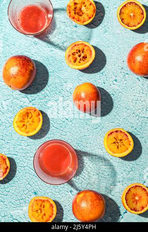 Hälften von reifen leckeren gepressten Blutorangen, die mit transparenten Gläsern roten Safts in einem hellen Raum mit Sonnenschein auf den Tisch gelegt wurden Stockfoto
