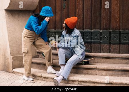 Optimistische Freundinnen surfen in den sozialen Medien auf dem Handy, während sie sich in der Nähe des Eingangs eines Wohnhauses in der Stadt angusten Stockfoto