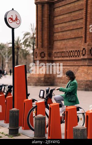 Seitenansicht einer Frau in grüner Jacke, die in der Nähe des Mietbahnhofs mit Zahlungsterminal auf der Straße der Stadt auf das Fahrrad fährt Stockfoto