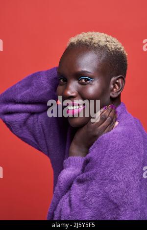 Nahaufnahme einer fröhlichen Afrikanerin mit kurzen Haaren in einer Studioaufnahme mit rotem Hintergrund Stockfoto