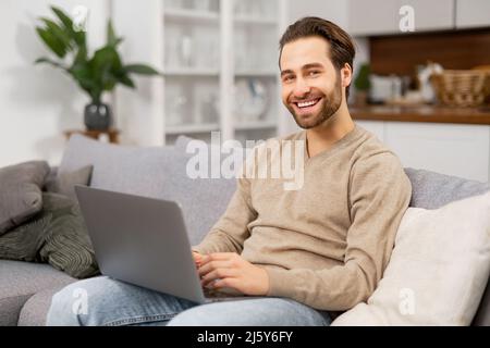 Lächelnder junger bärtiger Mann benutzt zu Hause einen Laptop. Typ mit Gerät, das auf der Couch sitzt, im Internet surft, Nachrichten ansieht, funktioniert von zu Hause aus Stockfoto