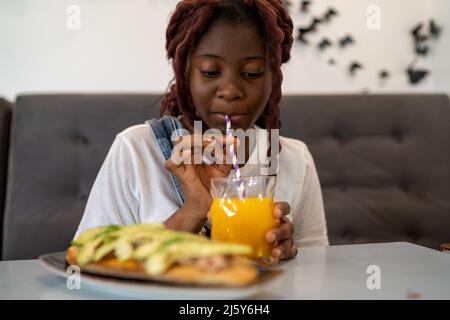 Junge afroamerikanische Frau trinkt frischen Orangensaft im Glas aus Stroh, während sie im hellen Café am Tisch sitzt Stockfoto