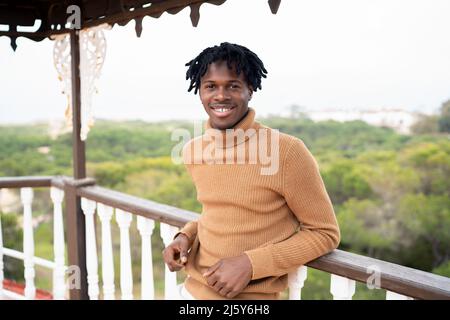 Fröhliches afroamerikanisches Männchen im Rollkragen, das auf die Kamera schaut, während es auf der Terrasse des Hauses mit einem Holzzaun auf dem Land steht Stockfoto