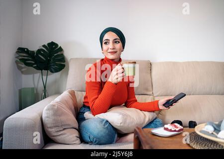 Trendy schöne afroamerikanische Frau mit lockigen Haaren und in blauem Kleid sitzen auf einem Stuhl im Studio und Blick auf die Kamera Stockfoto