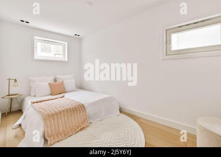 Komfortables weißes Bett mit Karomusterboden auf Teppich im hellen Schlafzimmer mit Pouf am Tisch und kleinem Fenster zu Hause Stockfoto