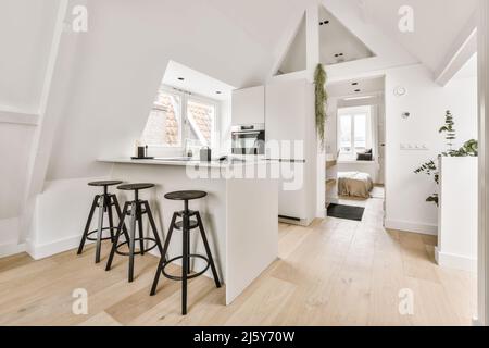 Barhocker an der Küchentheke mit weißen Schränken und modernen Geräten in einem hellen, geräumigen Apartment mit Topfpflanzen und Fenstern Stockfoto
