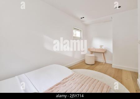 Komfortables weißes Bett mit Karomusterboden auf Teppich im hellen Schlafzimmer mit Pouf am Tisch und kleinem Fenster zu Hause Stockfoto