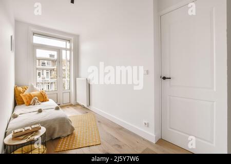 Komfortables Bett mit farbenfrohen Kissen und einem Stofftier, das in einem hellen Schlafzimmer mit weißen Wänden und Heizkörper neben dem Fenster platziert ist Stockfoto