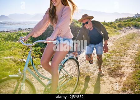 Happy Boyfriend schiebt Freundin auf Fahrrad auf sonnigen Strandweg Stockfoto