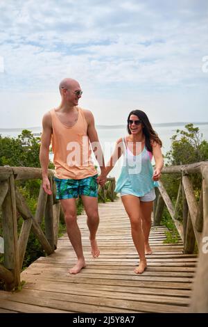 Glückliches Paar, das sich die Hände hält und auf der Strandpromenade läuft Stockfoto