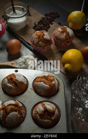 Blick von oben auf leckere, frisch gebackene Muffins, die auf dem Tisch serviert werden, mit Zitronen und Zucker in der Nähe von Schokoladentropfen in der hellen Küche Stockfoto