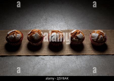 Blick von oben auf süße frisch gebackene Muffins mit Puderzucker, die in einer Reihe auf Textilien auf grauem Hintergrund in einer hellen Küche angeordnet sind Stockfoto