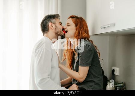 Seitenansicht eines liebenden Paares mit geschlossenen Augen, das sich mit Erdbeere küsst, während man Zeit zusammen in der hellen Küche zu Hause verbringt Stockfoto