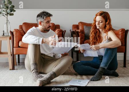 Ein ganzer Körper von ernsthaften Paaren, die Dokumente lesen, während sie zusammen auf dem Boden in der Nähe von Sesseln im hellen Wohnzimmer zu Hause sitzen Stockfoto