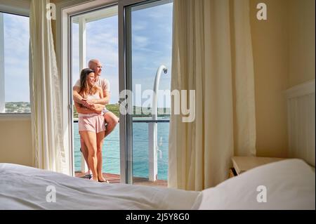 Ein glückliches Paar, das sich auf dem Hotelbalkon mit sonnigem Meerblick umarmte Stockfoto