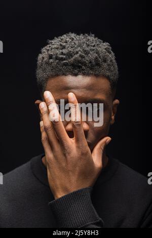 Schwarzer Mann mit kurzen lockigen Haaren, die die Hälfte des Gesichts mit Händen auf dunklem Hintergrund im Studio bedecken Stockfoto