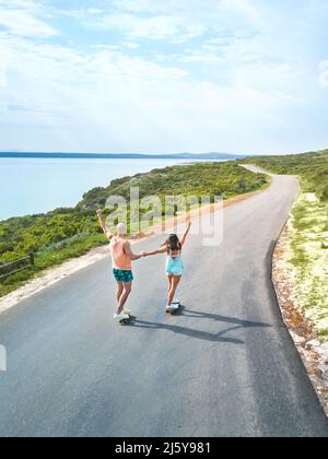 Sorgloses Paar, das auf der sonnigen Ocean Road Skateboards reitet Stockfoto