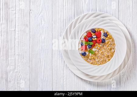 Geschälter Haferbrei mit Vollkornkorn und frischen Himbeeren, Heidelbeeren und Kokosnusssplittern in einer weißen Schüssel auf dem Holztisch, horizontale Ansicht von oben, Stockfoto