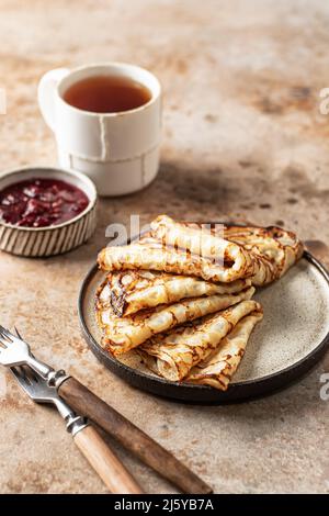 Crepes in brauner Platte. Frühstück Zusammensetzung mit Blinis, rustikalen Gabeln, Tee, Marmelade. Vertikale Ausrichtung Stockfoto