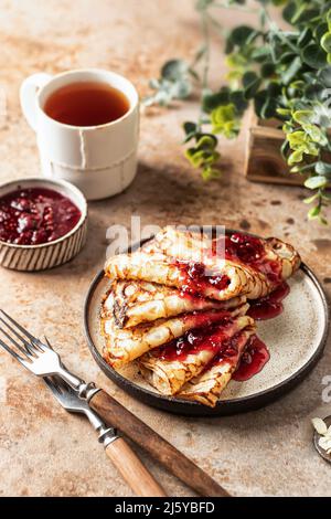 Crepes mit Himbeermarmelade in Keramikplatte auf braunem Hintergrund. Frühstück Zusammensetzung mit Lebensmitteln, Gabeln, Tee und Gemüse im Hintergrund Stockfoto