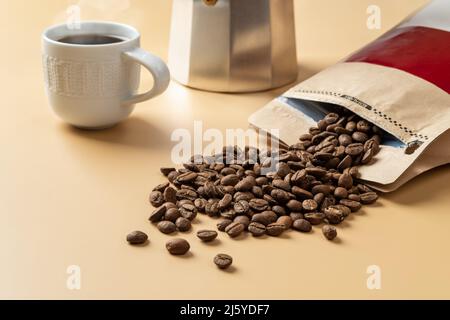 Aus einem braunen Papierbeutel auf beigem Hintergrund verschüttete Bio-Kaffeebohnen. Ganze geröstete Kaffeebohnen, eine Tasse heißen Kaffee und eine Geysir-Kaffeemaschine. Stockfoto