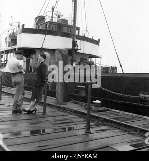 1960, historisch, stehend auf einem feuchten Holzkai, ein junger Mann, der in T-Shirt und Mütze mit einer jungen Frau spricht, Birkenhead Docks, Mersey, Liverpool, England, VEREINIGTES KÖNIGREICH. Das Frachtschiff Seatern ist vertäut. Stockfoto