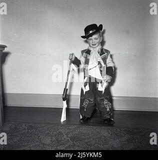 1960s, historisch, in einem Raum, ein kleiner Junge, der in seinem Cowboy-Kostüm für ein Foto steht, einen wilden Westernhut mit „Kentucky Kid“ auf dem Kopf trägt und ein „Davy Crocket“-Spielzeuggewehr in der Hand hält, England, Großbritannien. Davy Crockett, ein amerikanischer Volksheld, der 1836 im Almo, Texas, kämpfte und starb, ist bekannt als der „König der wilden Grenze“. Stockfoto