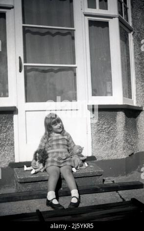 1960s, historisch, einfach nur chillen... im Sommer und ein junges Mädchen, das mit ihren zwei Puppen auf den Stufen vor der Hintertür ihres Hauses, England, Großbritannien, sitzt. Stockfoto