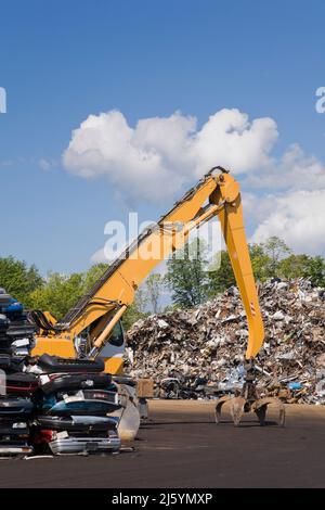 Kranlader mit Greifarm und Klauenklemme vor dem Stapel ausrangierter Autos, metallischer Haushalts- und Industriegegenstände auf dem Schrottplatz. Stockfoto