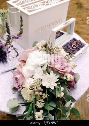 Lavendelhochzeit. Eheringe mit violetten Blumenverzierungen auf weißem Tisch Stockfoto
