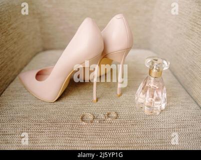 Goldenes Hochzeitsband und Verlobungsring, Parfümflasche und pudrige Schuhe in Staubrosa. Nahaufnahme, flach liegend Stockfoto
