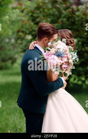 Hochzeitspaar küsst sich im grünen Park. Schöner Hochzeitsstrauß mit weißen, rosa, blauen Rosen. Glückliche Braut und Bräutigam. Sommerhochzeit, Brautpaar Stockfoto