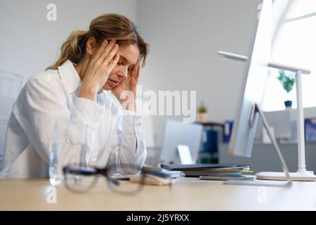 Müde Geschäftsfrau mit Kopfschmerzen am Schreibtisch. Porträt einer erschöpften Büroarbeiterin Stockfoto