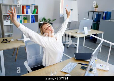 Die reife Geschäftsfrau streckt sich, während sie nach einer sitzenden Arbeit an ihrem Schreibtisch im Büro sitzt Stockfoto